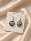A pair of dangling earrings with colorful circular mandala designs in silver hooks, displayed on a white card over a beige satin fabric background.