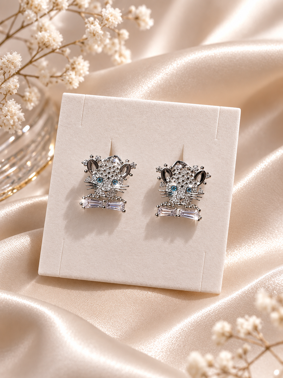 A pair of silver-toned stud earrings with clear crystal details and light blue center stones, displayed on a cream jewelry card over draped champagne satin fabric with small white flowers.