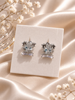 A pair of silver-toned stud earrings with clear crystal details and light blue center stones, displayed on a cream jewelry card over draped champagne satin fabric with small white flowers.
