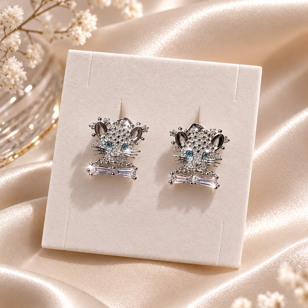 A pair of silver-toned stud earrings with clear crystal details and light blue center stones, displayed on a cream jewelry card over draped champagne satin fabric with small white flowers.
