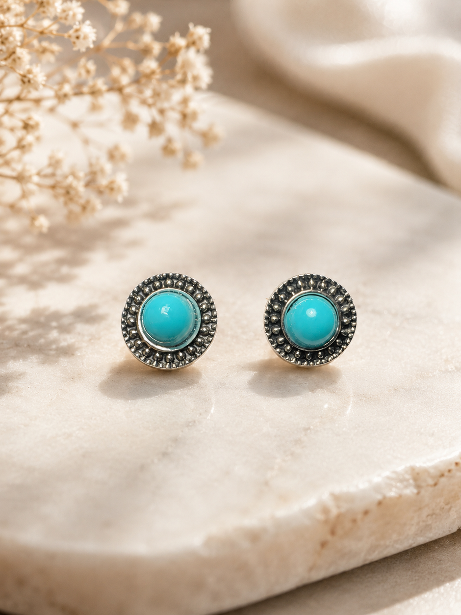 A pair of round stud earrings with bright turquoise centers and dark silver-toned textured halos, displayed on a light beige stone surface with dried flowers in the background.