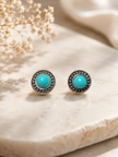 A pair of round stud earrings with bright turquoise centers and dark silver-toned textured halos, displayed on a light beige stone surface with dried flowers in the background.