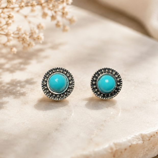 A pair of round stud earrings with bright turquoise centers and dark silver-toned textured halos, displayed on a light beige stone surface with dried flowers in the background.