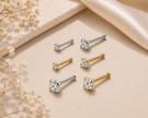 Six stud earrings arranged on a beige surface with decorative dried flowers and satin fabric. The earrings include silver and gold posts with clear gemstones in round and square settings.