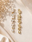 Two long drop earrings displayed on a warm cream surface. One earring features clear teardrop-shaped crystals, and the other features textured gold-toned crystal leaves.