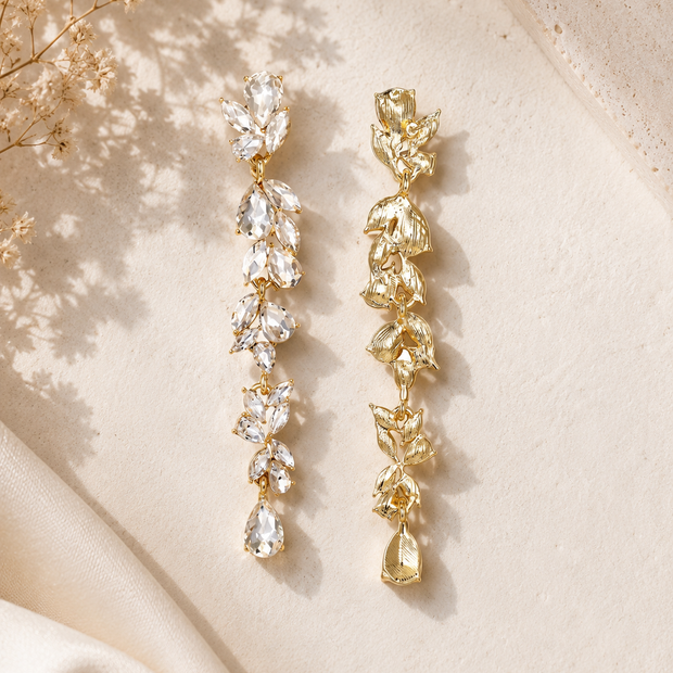 Two long drop earrings displayed on a warm cream surface. One earring features clear teardrop-shaped crystals, and the other features textured gold-toned crystal leaves.