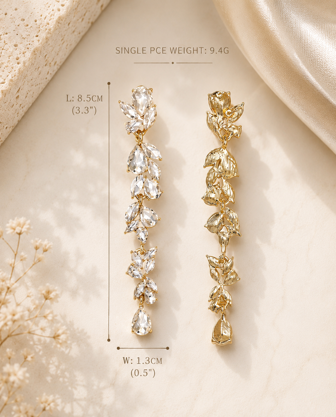 Two long drop earrings displayed on a light beige surface with decorative fabric and dried flowers. One earring is clear crystal and the other is gold-tone, with measurement text and dimension lines showing length and width.
