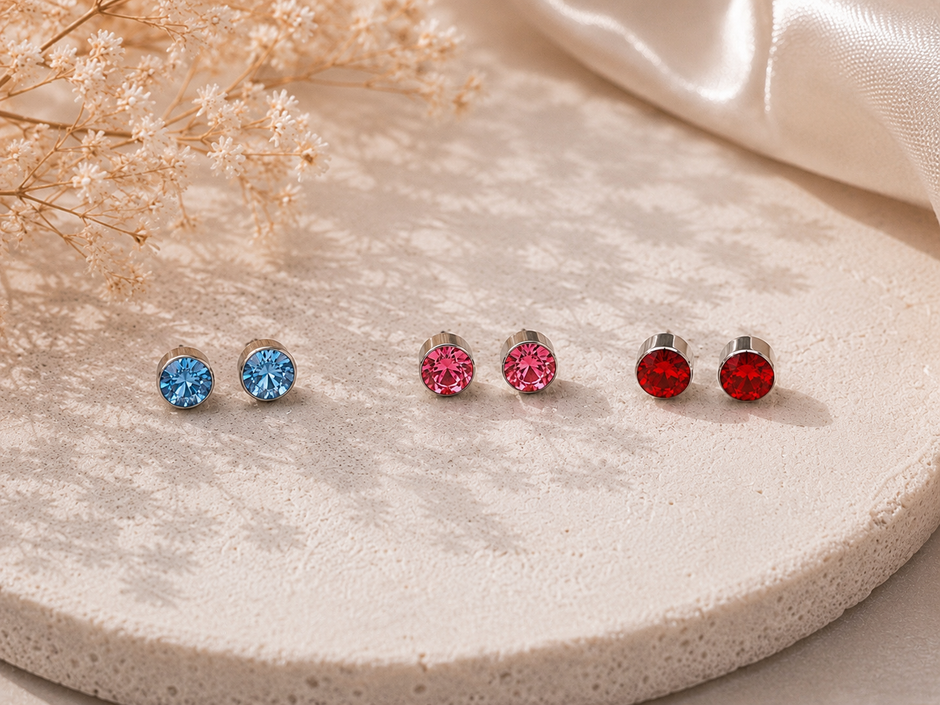 Three pairs of round stud earrings in blue, pink, and red, displayed on a textured beige stone tray with dried flowers and a satin ribbon.