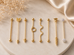 Seven gold-toned lip barbell piercings in different decorative tip styles are arranged on a round beige stone slab with soft satin fabric and dried flowers in the background.