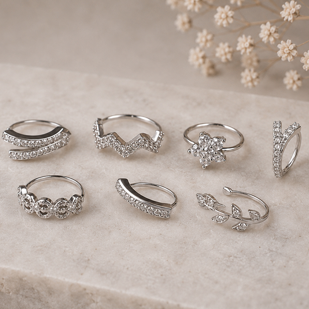 Seven silver-toned rings with clear stones in various organic and floral shapes, arranged on a light beige marble slab with dried baby's breath flowers in the background.