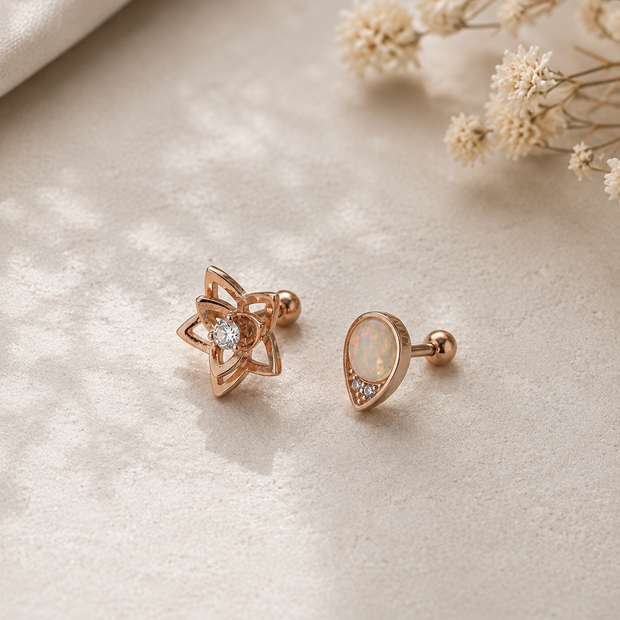 A pair of rose gold stud earrings, one in the shape of a five-pointed star with a clear central gemstone, and the other a teardrop-shaped stud with an iridescent white opal-like center and a small clear stone, displayed on a textured beige fabric with dried white flowers in the background.