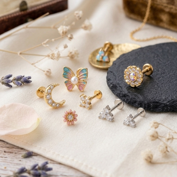 A styled flat-lay of several small gold-toned earrings and a butterfly stud arranged on cream fabric with dried flowers, lavender sprigs, a dark stone slab, and a wooden box in the background.