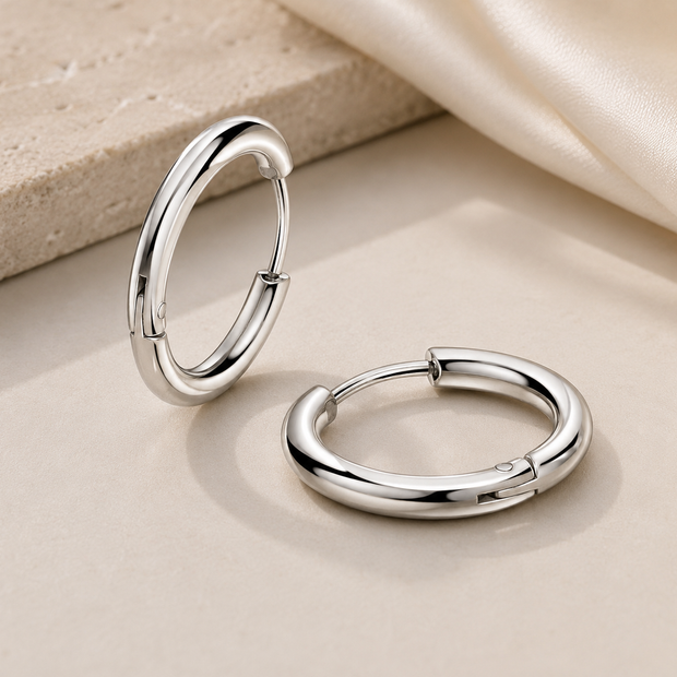 Two polished silver hoop earrings displayed on a beige surface with a textured stone slab and draped satin fabric in the background.