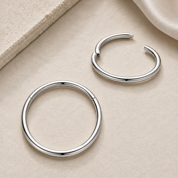 Two polished silver hoop earrings displayed on a textured beige surface with a soft cream fabric corner in the background.