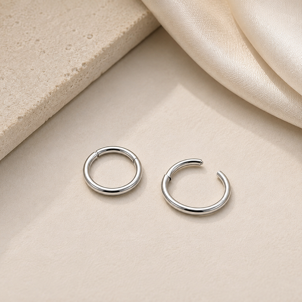 A pair of polished silver-tone open hoop earrings resting on a light beige textured surface with a cream satin fabric draped in the upper right corner.