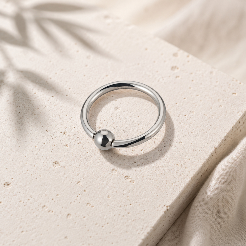 A polished silver ring with a small spherical bead detail, resting on a textured beige stone slab with soft fabric and leaf shadows in the background.