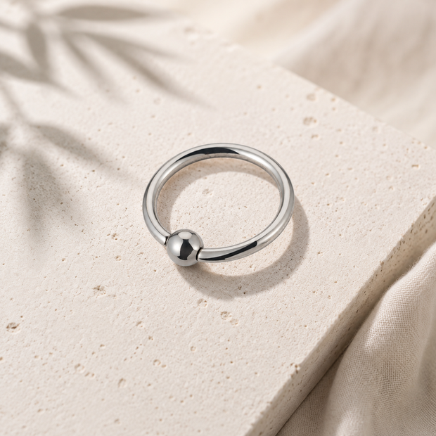 A polished silver ring with a small spherical bead detail, resting on a textured beige stone slab with soft fabric and leaf shadows in the background.