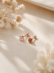 A pair of rose gold stud earrings, one shaped like a small heart with a clear crystal and the other as a rose, displayed on a beige satin fabric with dried flowers.