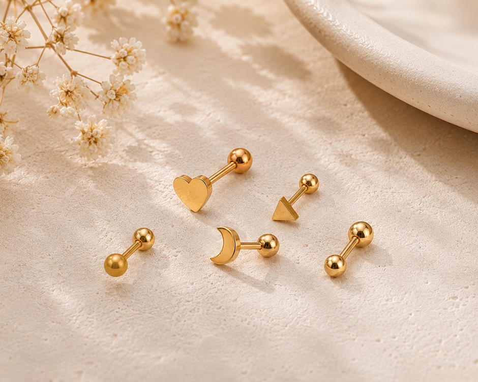 Five gold-colored labret stud earrings with different minimalist designs (heart, triangle, and three geometric shapes) arranged on a textured beige surface with soft shadows and dried baby's breath flowers.