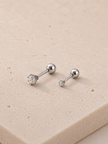A pair of silver-toned labret stud earrings with round bezel-set clear gemstones, displayed on a textured beige stone surface.