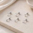 Six silver-toned labret stud earrings with different minimalist backings, including a heart, crescent moon, and triangle shapes, arranged on a textured beige stone slab.