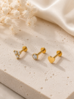 Three gold-colored stud earrings displayed on a textured beige stone slab with white baby's breath flowers and a cream satin fabric in the background. The earrings include a heart-shaped stud, a round clear gemstone stud, and a small clear gemstone cluster stud.