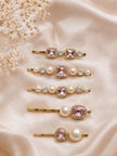 Six gold hair claw clips styled on a beige satin fabric background with dried baby's breath flowers. The clips feature pink gemstones, faux pearls, and clear rhinestones.
