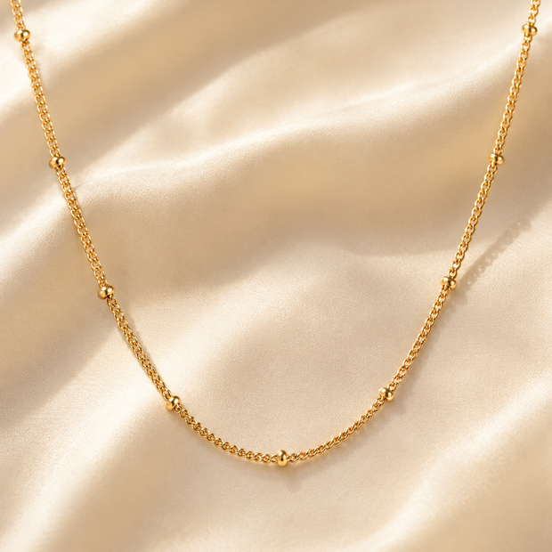 A delicate gold chain necklace with small bead accents, draped elegantly on a soft, cream-colored satin fabric background.