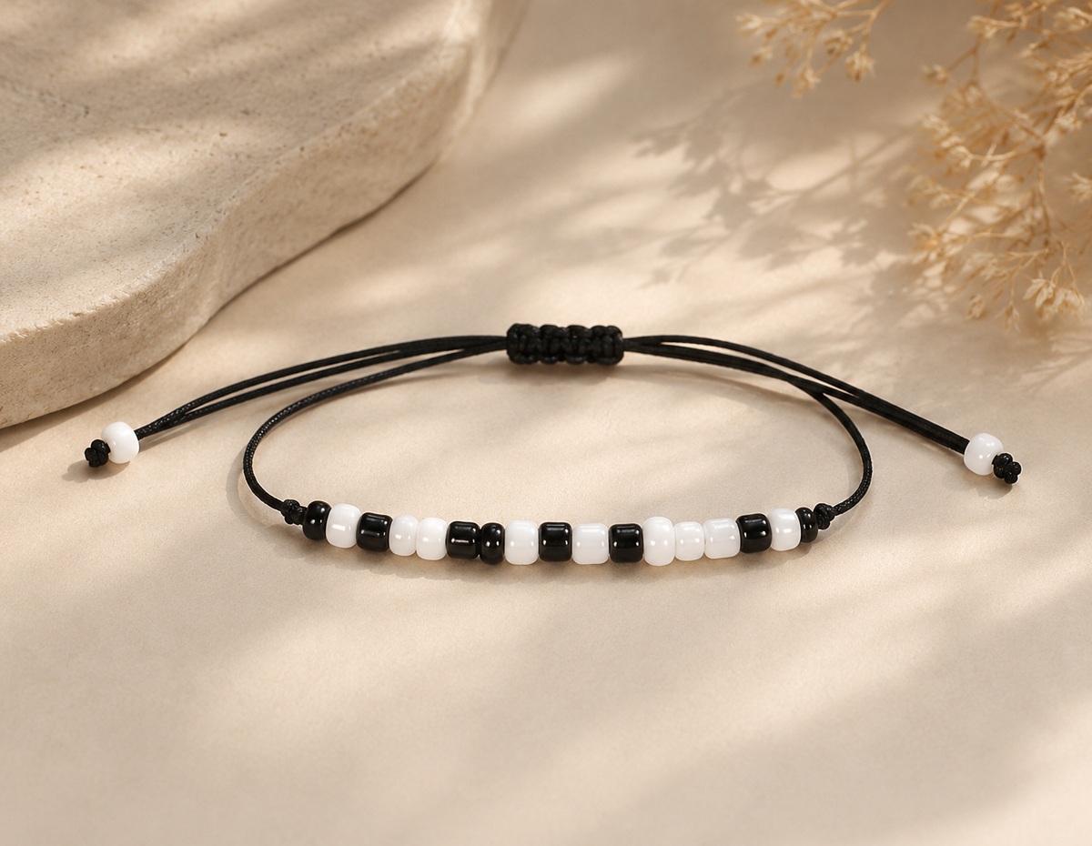 A beaded adjustable cord bracelet with alternating black and white beads, displayed on a warm beige surface with soft shadows and dried floral decor.