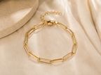A gold paperclip chain bracelet with an adjustable lobster clasp, displayed on draped cream satin fabric next to a textured stone prop.