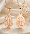 Two rose gold Miraculous Medal necklaces displayed side-by-side on a light beige stone surface with a soft white fabric backdrop and dried flowers. One necklace shows the front of the medal with an embossed figure of the Virgin Mary, and the other shows the back with a cross, monogram, and stars.