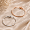 Two snake-style bangles, one rose gold and one silver, displayed on a draped beige satin fabric with dried flowers in the corner.