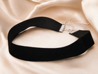A black velvet choker necklace with a silver-tone metal clasp and chain extender, displayed on a draped beige satin fabric background.