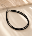 A black multi-strand choker necklace with a silver clasp and extension chain, styled on a warm cream fabric with soft diagonal shadows.