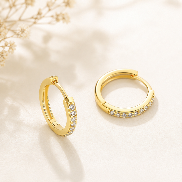 A pair of gold huggie hoop earrings with small clear pavé stones along the front, displayed on a soft cream surface with a delicate dried floral prop in the background.
