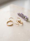 A pair of gold crescent-shaped hoop earrings displayed on a light beige stone surface next to a sprig of purple lavender.