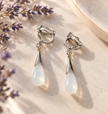 A pair of silver-toned drop earrings with teardrop-shaped milky white pendants, displayed on a light beige stone surface with dried lavender sprigs.