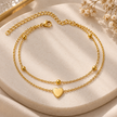 A double-layered gold chain anklet with a polished heart charm and small gold beads, displayed on a round beige stone tray with dried flowers.