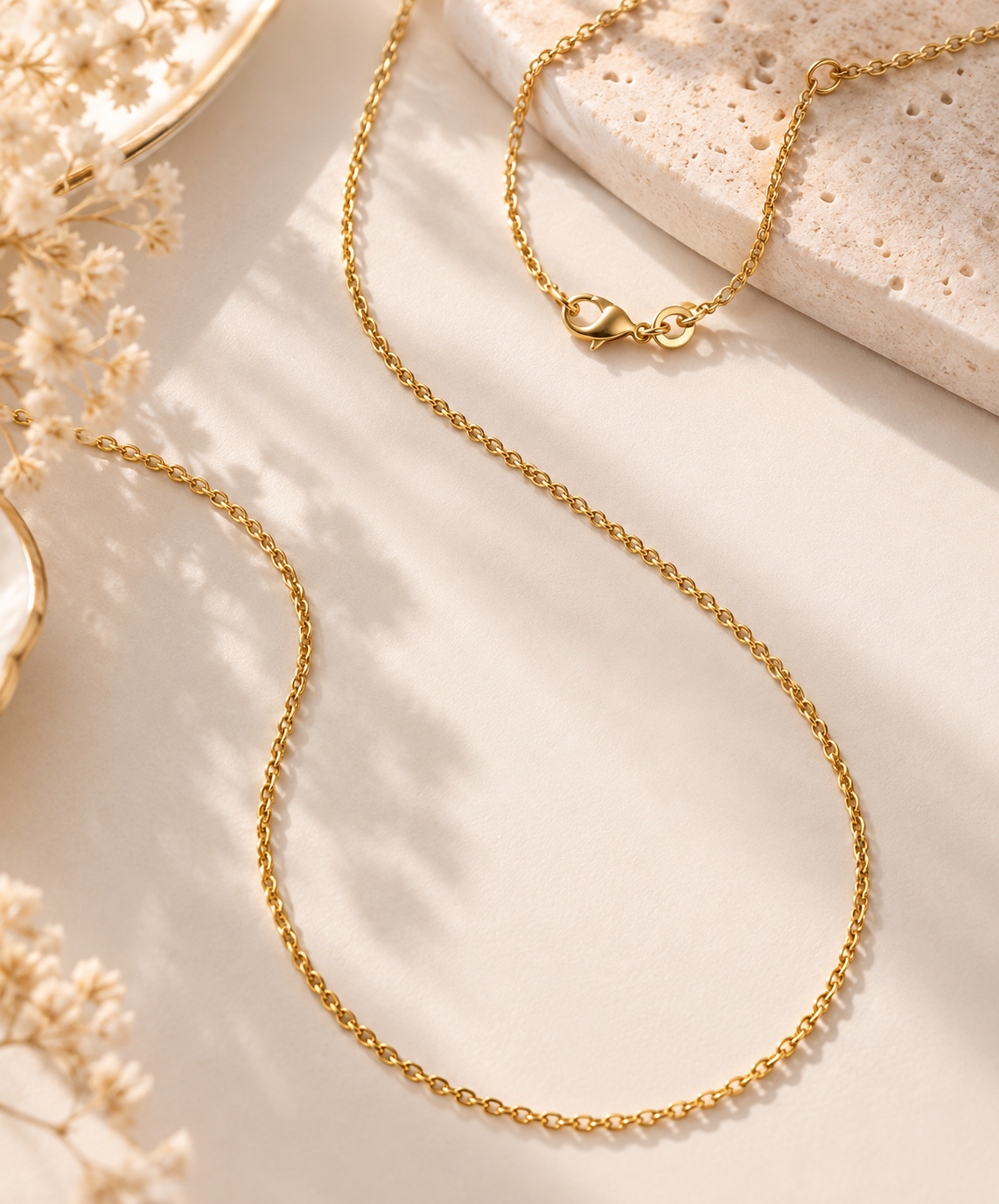 A delicate gold chain necklace with a lobster clasp, styled in a flat-lay composition on a warm beige surface with dried flowers and a textured stone slab.