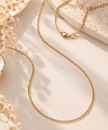 A delicate gold chain necklace with a lobster clasp, styled in a flat-lay composition on a warm beige surface with dried flowers and a textured stone slab.