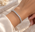 A close-up of a silver tennis bracelet with clear stones worn on a woman's wrist, styled with a white satin sleeve and a decorative book in the background.