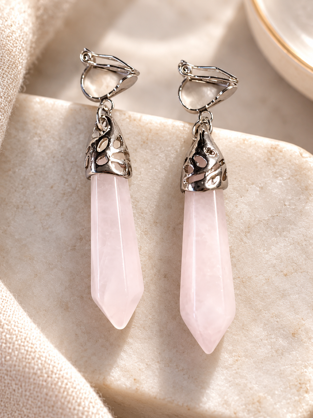 A pair of silver-toned dangle earrings with elongated pink crystal points, displayed on a textured beige stone surface with soft natural light.