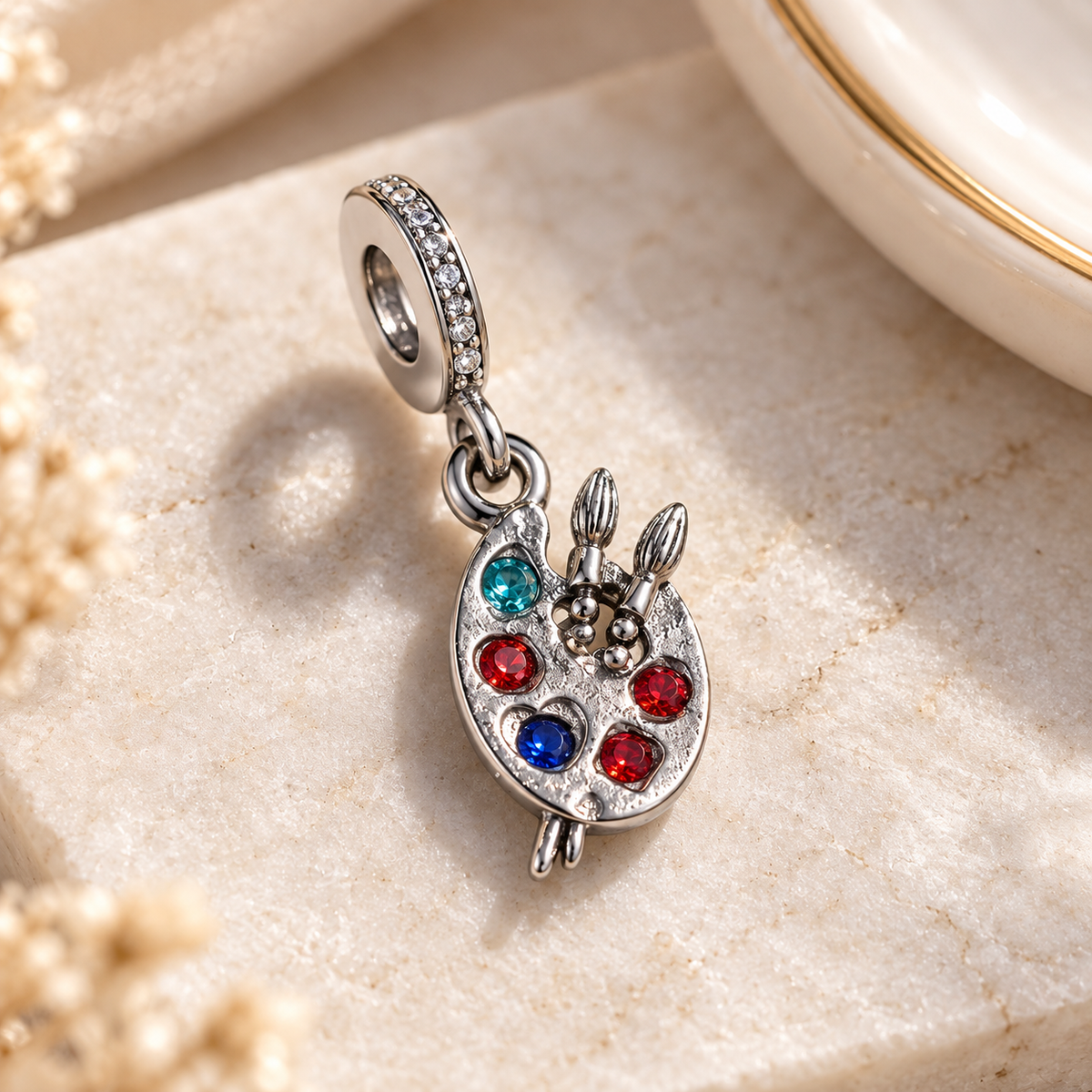 A silver charm pendant shaped like a paint palette with colorful rhinestones, photographed on a light stone surface.