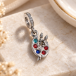 A silver charm pendant shaped like a paint palette with colorful rhinestones, photographed on a light stone surface.