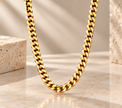 A close-up of a thick gold Cuban link chain necklace draped over a light marble surface, with a warm beige background and decorative stone blocks on either side.