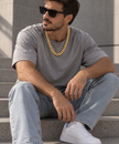 A man sitting on concrete steps wearing a thick gold chain necklace, a gray t-shirt, light-wash blue jeans, and white sneakers.