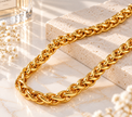 A chunky gold chain necklace with a braided, woven link design, styled on a light marble surface with a textured stone block and small white flowers.