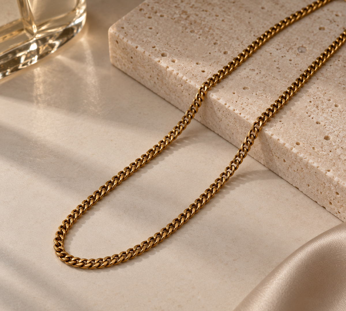 A gold-toned metal chain necklace draped over a light beige stone slab and satin fabric, with part of a glass visible in the upper left corner.