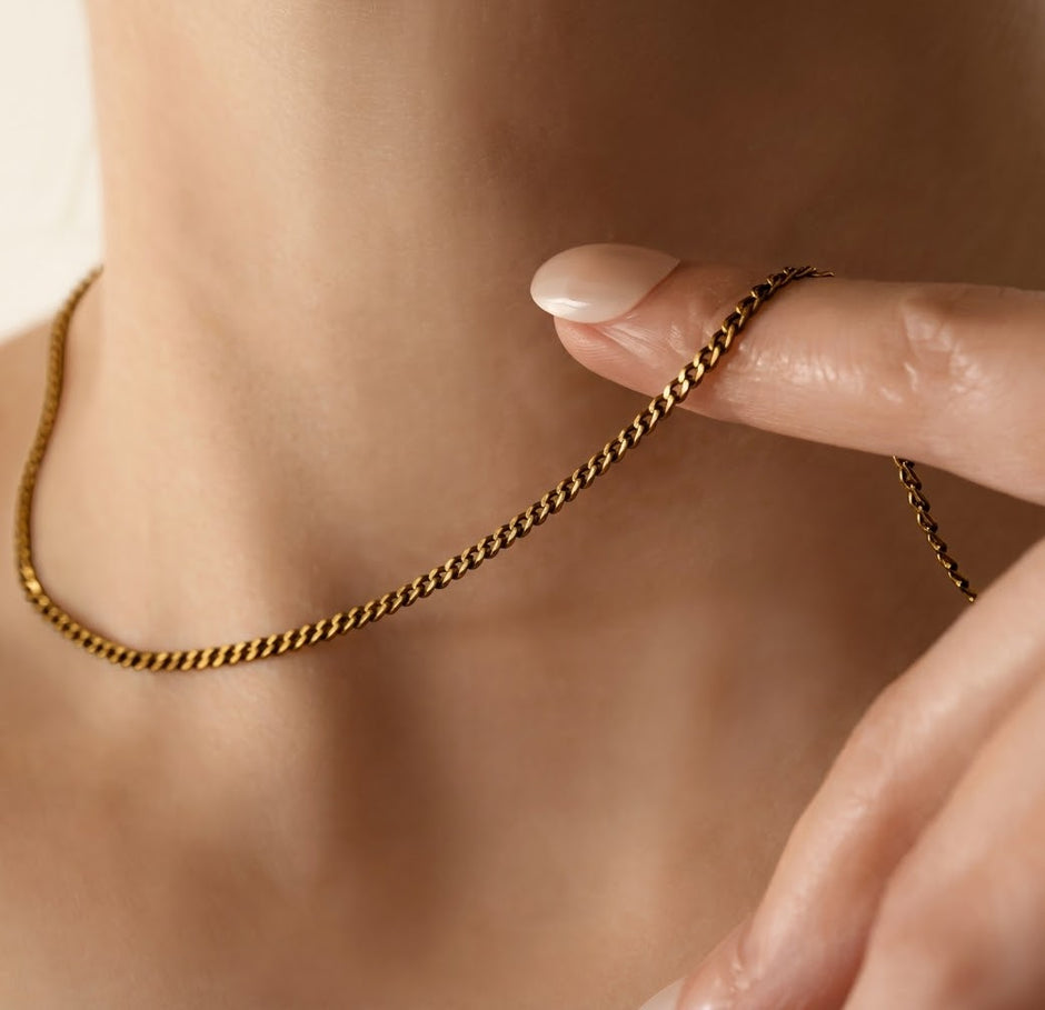 A close-up of a delicate gold chain necklace worn around a person's neck, with a hand gently touching the chain near the collarbone.