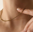A close-up of a delicate gold chain necklace worn around a person's neck, with a hand gently touching the chain near the collarbone.